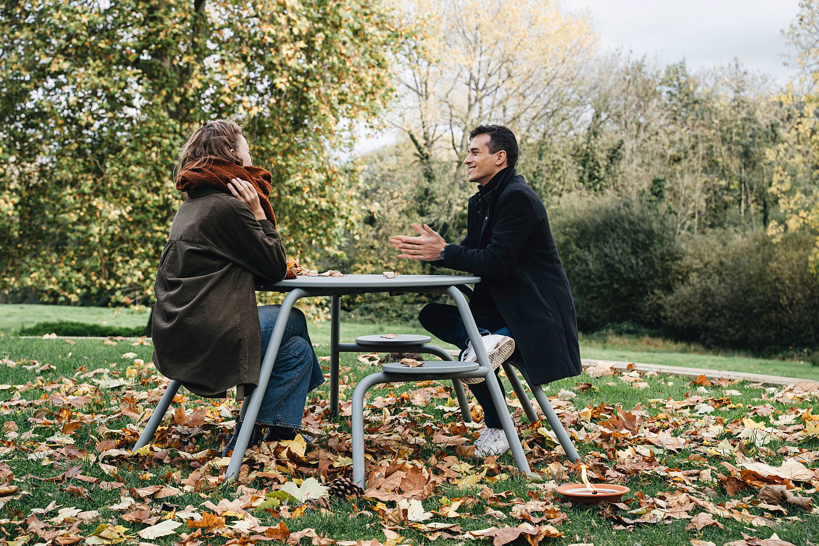 Een man en vrouw praten aan een grijze Virus picknicktafel in een park vol herfstbladeren.
