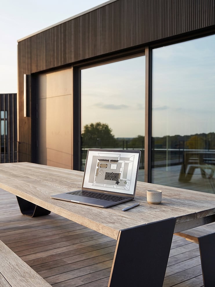 Laptop displaying interior floor-plan software on a wooden outdoor table, with a coffee cup and pen beside it.