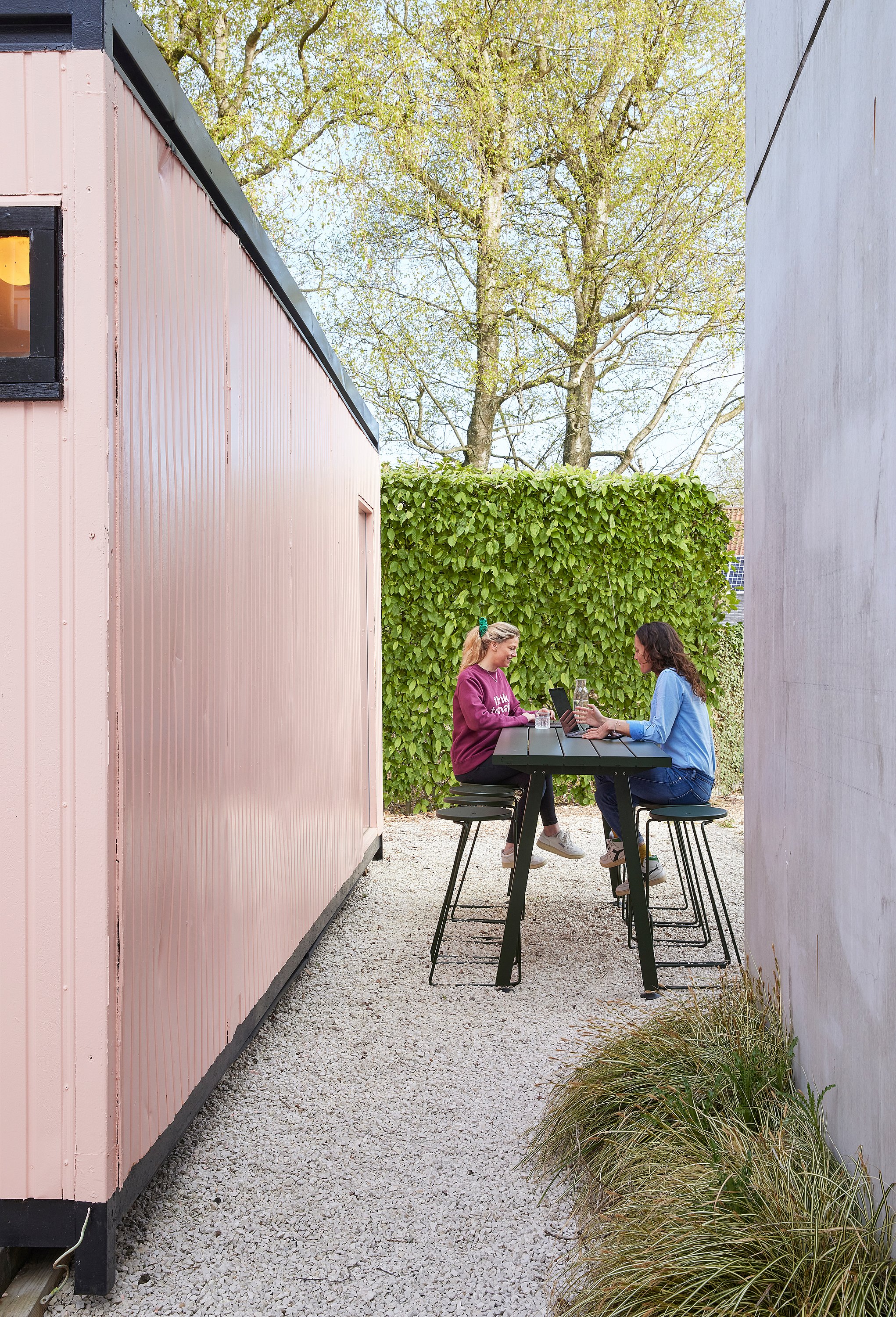 Two women working on laptops at a dark green Marina table with matching stools outside a pink garden office pod.
