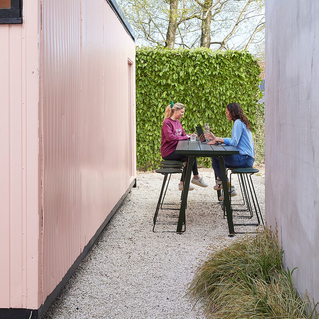 Zwei Frauen arbeiten an Laptops an einem dunkelgrünen Marina-Tisch mit passenden Hockern vor einem rosa Gartenbüro.