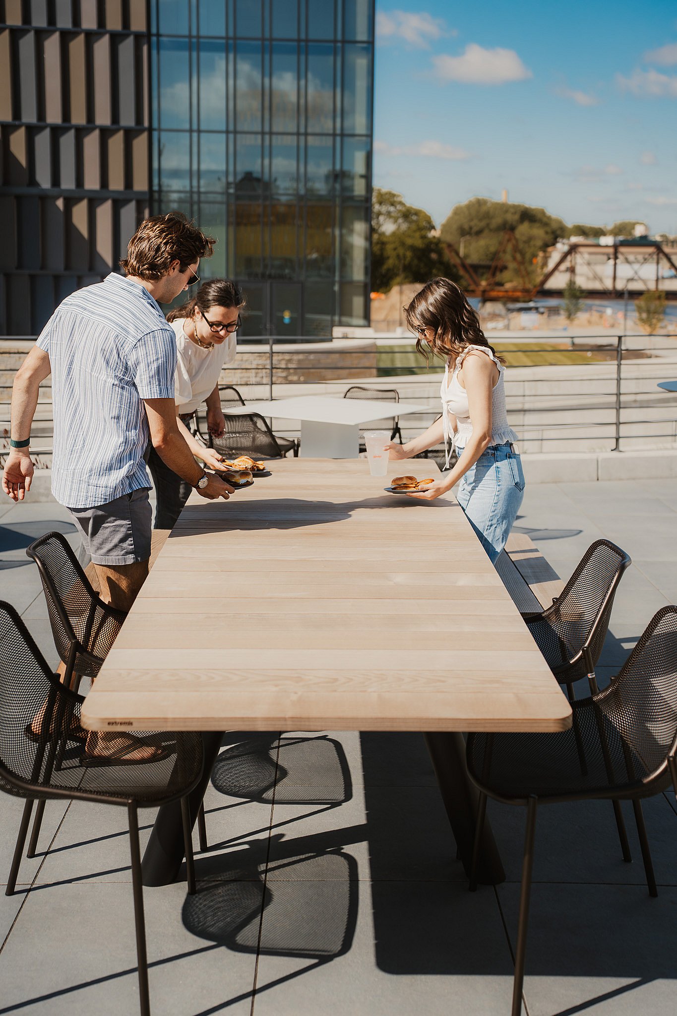 Guests gathering at an Extremis Panigiri outdoor dining table beside the glass facade of Acrisure Amphitheater