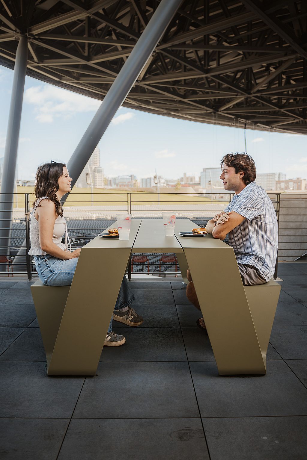 Guests at an Extremis Hopper aluminum picnic table under the canopy, with the Grand Rapids skyline behind